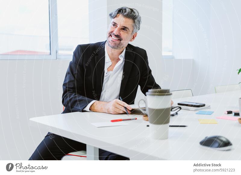 An affable executive in a black suit and white shirt sits at his office desk, smiling while taking notes, surrounded by work essentials cheerful professional