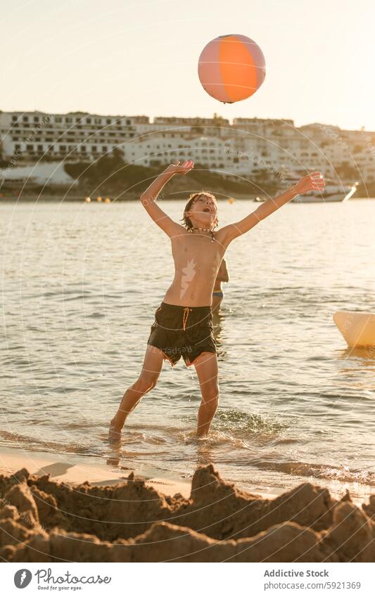 Happy boy playing with ball at shore throw sea playful vacation summer stand arms raised water beach building full body carefree happy freedom holiday lifestyle