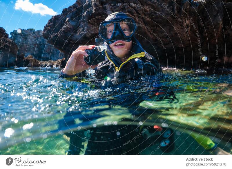 Portrait of anonymous male teen scuba diver wearing wetsuit and mask looking at camera while swimming in ocean against rock formations during summer vacation