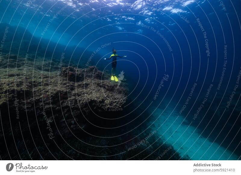 Full body of unrecognizable active teen boy wearing yellow flippers snorkeling by coral reef in deep ocean with crystal clear waters near rocky formation at Menorca