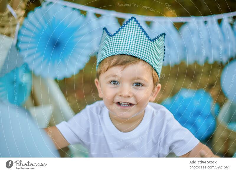 Toddler celebrating with balloons and decorations outside toddler boy smile celebration crown checkered outdoor blue white party happy cute child joy cheerful