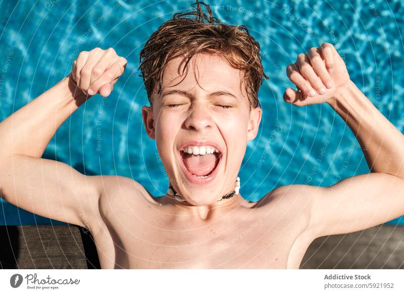 Excited Boy Flexing Muscles by the Pool on a Sunny Day boy child excitement pool swimming water sunny outdoors summer happiness wet hair shell necklace cheerful