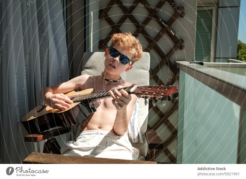 Young boy enjoys playing guitar in the sunlight music instrument child musician practice acoustic balcony outdoor leisure activity summer sunglasses relaxation