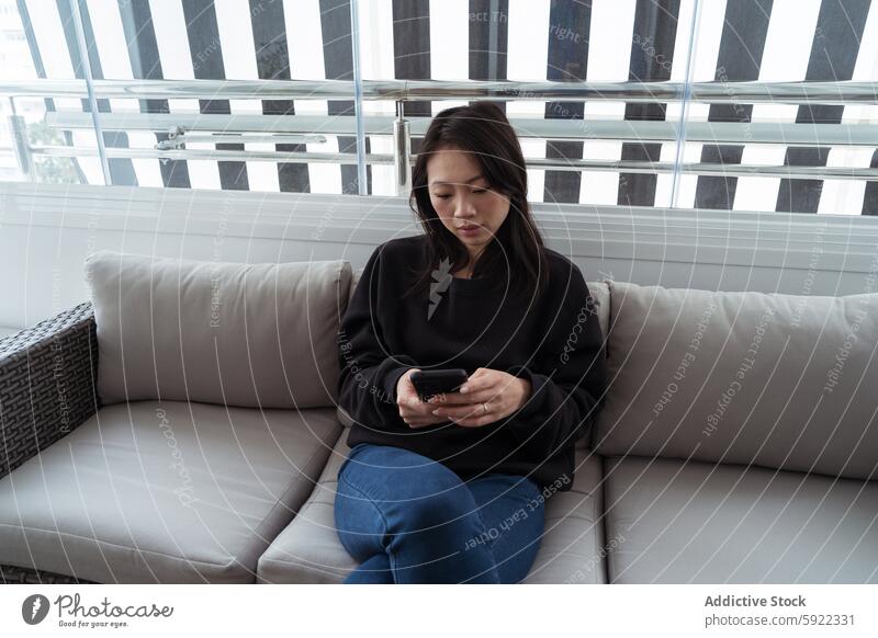 Young woman browsing on smartphone while seated on couch young adult mobile device sitting indoor window pattern modern focus comfortable casual screen