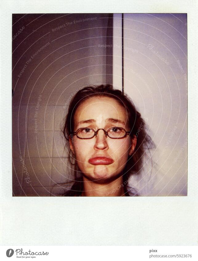 Polaroid of a woman in her early 20s with small glasses grimacing in front of a bathroom wall Grimace Eyeglasses Mouth Lips Sulk color cast Square square format