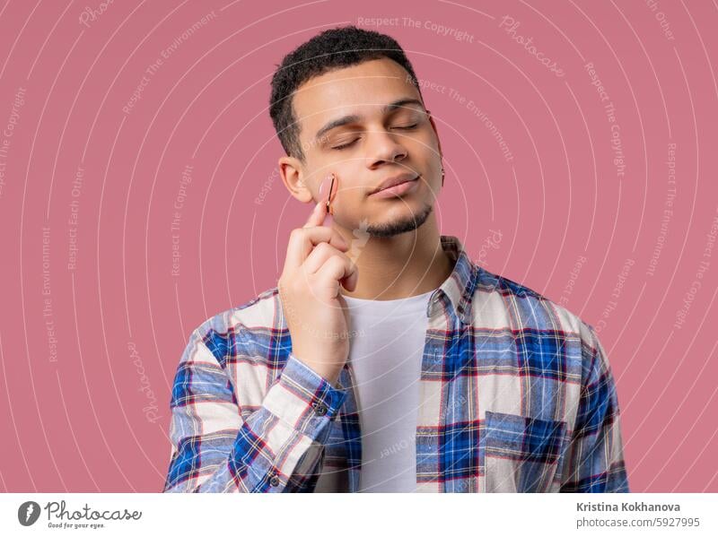 Young man with rose quartz stone roller on pink background. Facial self care adult anti aging attractive beautiful beauty body care clean cosmetic cosmetology