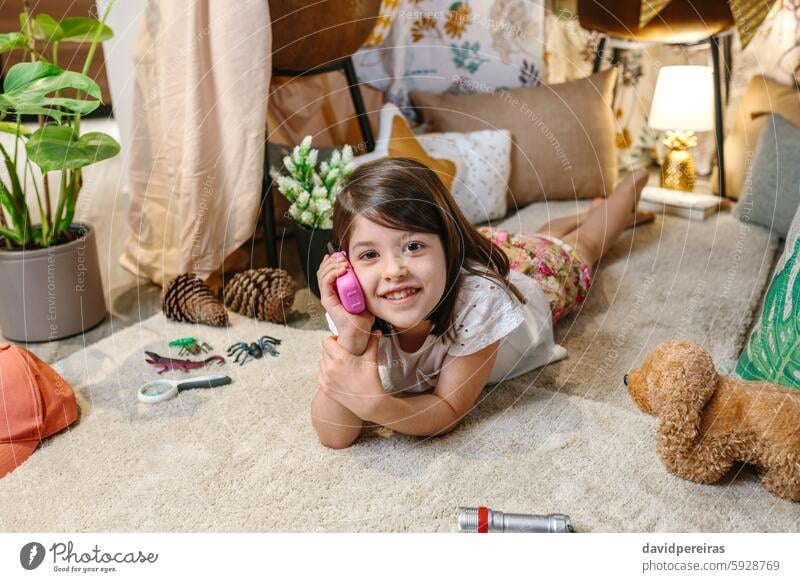 Smiling little girl looking at camera while using walkie-talkie lying over rug surrounded by cushion smile talking happy playing portrait child holding carpet