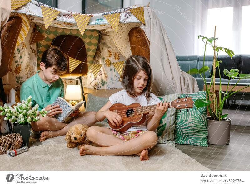 Little girl playing ukulele while boy reading book on handmade teepee in living room at home guitar children having fun shelter tent diy vacation camping house