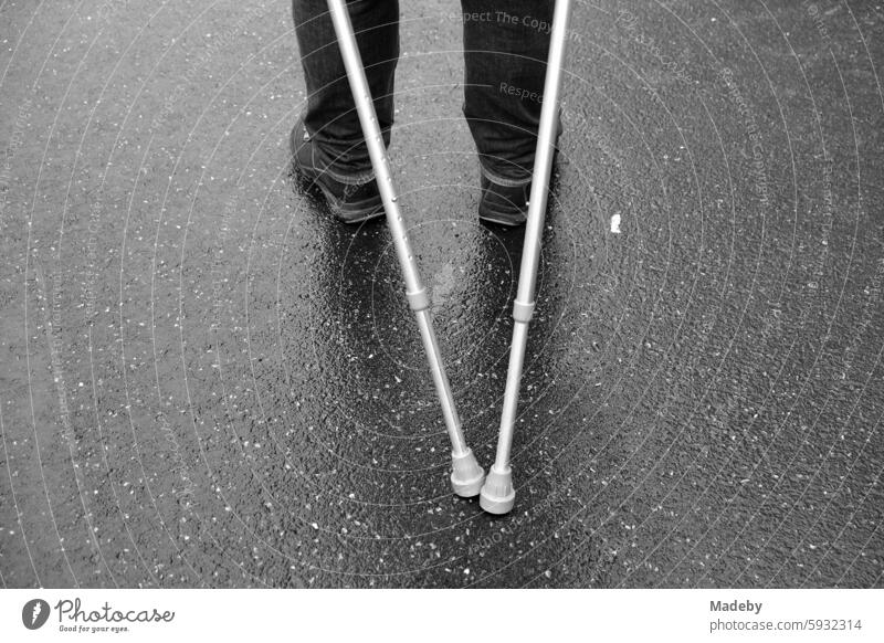 Man with crutches or walking sticks leans back and supports himself on wet gray asphalt at the Golden Oldies classic car meeting in Wettenberg Krofdorf-Gleiberg near Giessen in Hesse in black and white