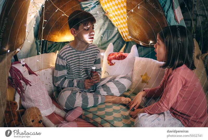 Boy illuminated with lantern telling a terror story to surprised girl sitting in dark living room boy tale children flashlight having fun young little homemade