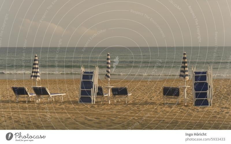 Unused blue and white beach loungers, partly stacked, and 3 folded umbrellas on a sandy beach. Behind them the Mediterranean Sea with moderate swell, a deserted beach, deserted, the photo is very symmetrical.