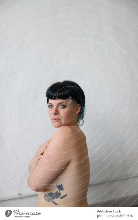 Young woman with tattoo looks at camera over shoulder, neutral background portrait white background gaze thoughtful expression female bangs dark hair