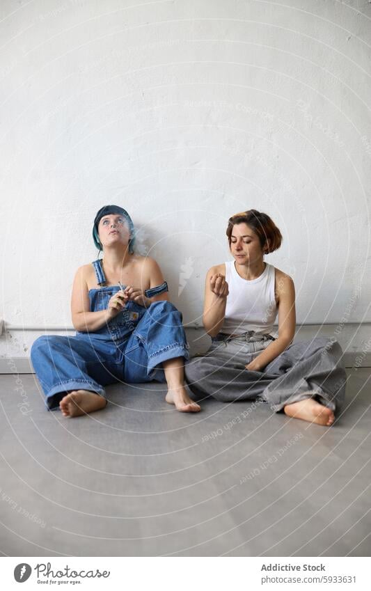 Two sisters sitting on floor, one looking away, other gazing down portrait seated wall blue hair brown hair denim top barefoot thoughtful pensive female emotion