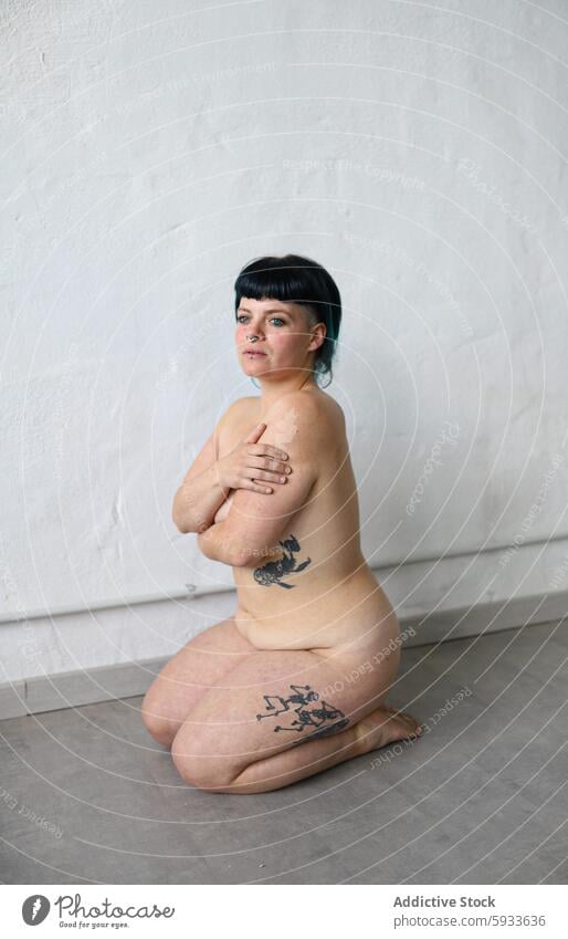 Woman with tattoos sitting against a plain backdrop woman portrait expression neutral indoor black hair bob cut skin tattoo art pose body positivity ink decor