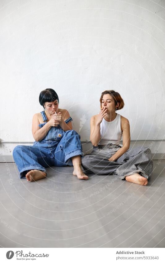 Two sisters in casual wear sitting and interacting adult denim interaction cigarette wall white floor grey contemplative relaxed portrait gaze away smoke