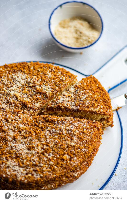 Top view of sliced homemade honey cake with toppings dessert sweet bake pastry crushed nuts plate blue rimmed ground nuts bowl white texture nutty crumble sugar