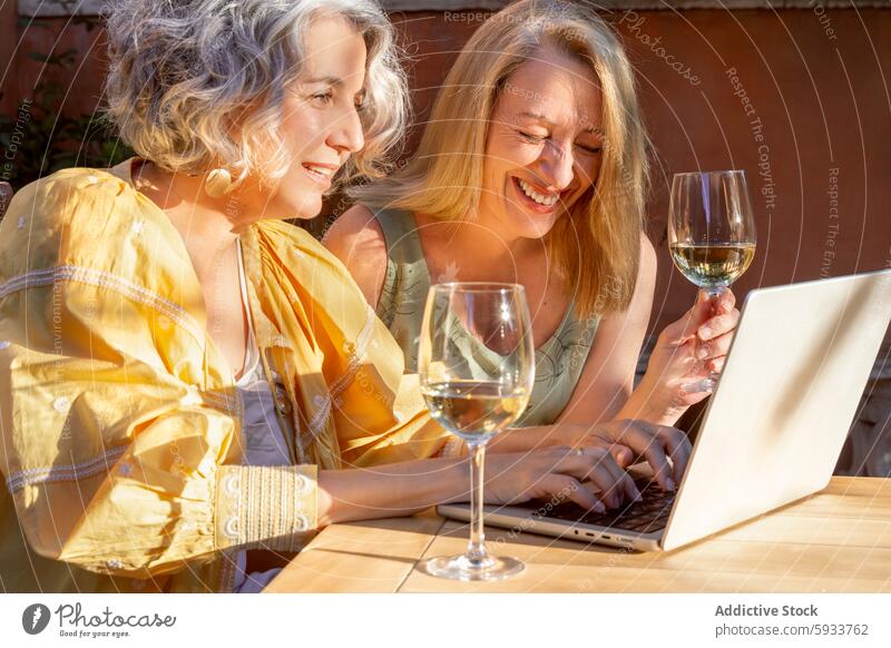 Two women enjoying wine while using laptop outdoors smiling middle-aged leisure friendship moment together technology internet garden summer sunshine cheerful