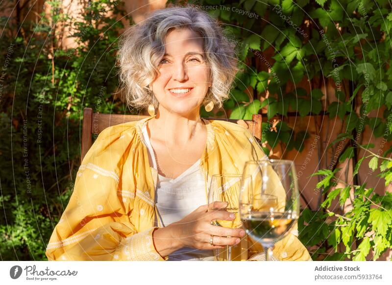 Mature woman enjoying wine in a garden setting mature glass outdoor sunlight smile looking at camera joyful sitting summer leisure relaxation foliage vine