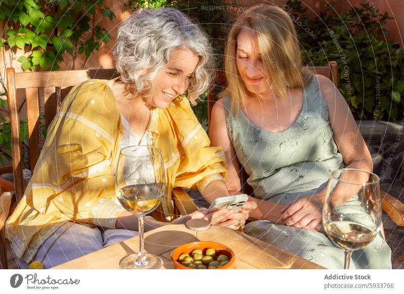 Two women enjoying a leisurely afternoon with wine outdoors mature senior friendship drinking garden table glass summer smartphone smiling enjoyment bonding