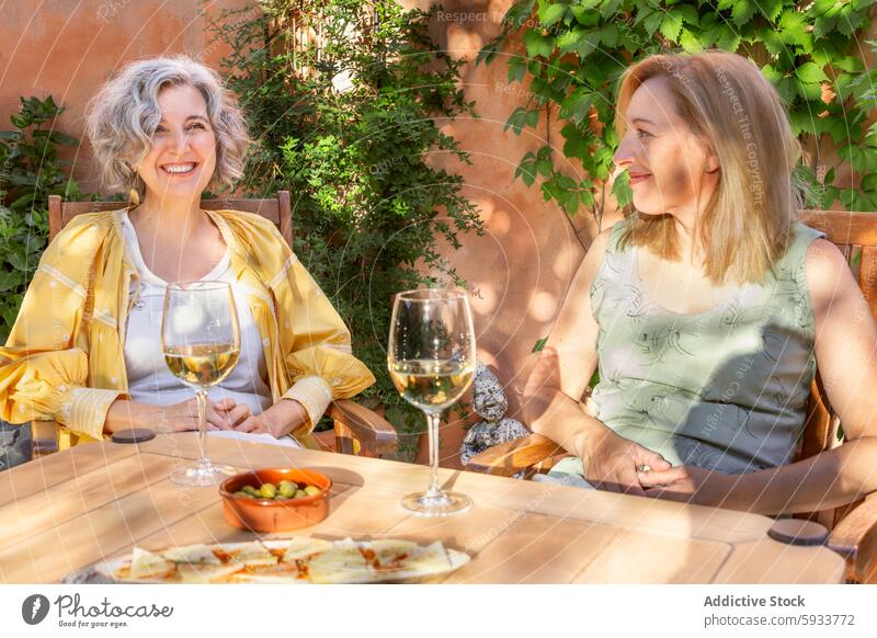 Two women enjoying wine and conversation outdoors smiling sunny table chair garden glass leisure enjoyment friendship vine summer daytime peaceful relaxing