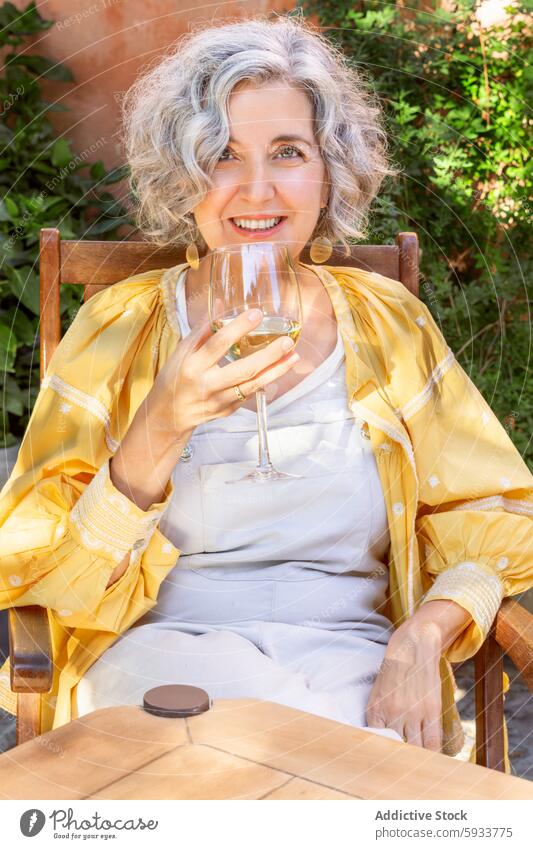 Woman enjoying wine outdoors in a relaxed setting woman leisure smile glass sit table relaxation happy middle-aged jacket white outfit yellow bright