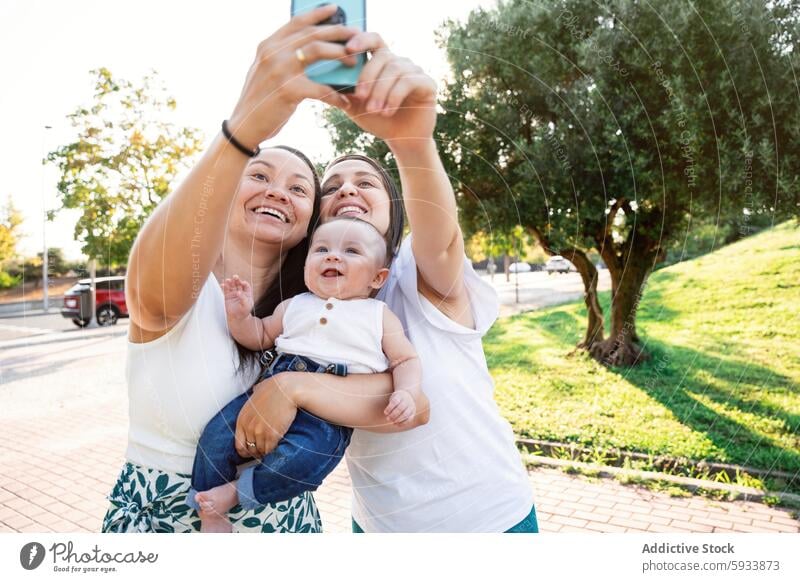 Lesbian couple taking selfie with their young son outdoors lesbian baby family smartphone joy happiness love parent woman summer sunlight park casual clothing