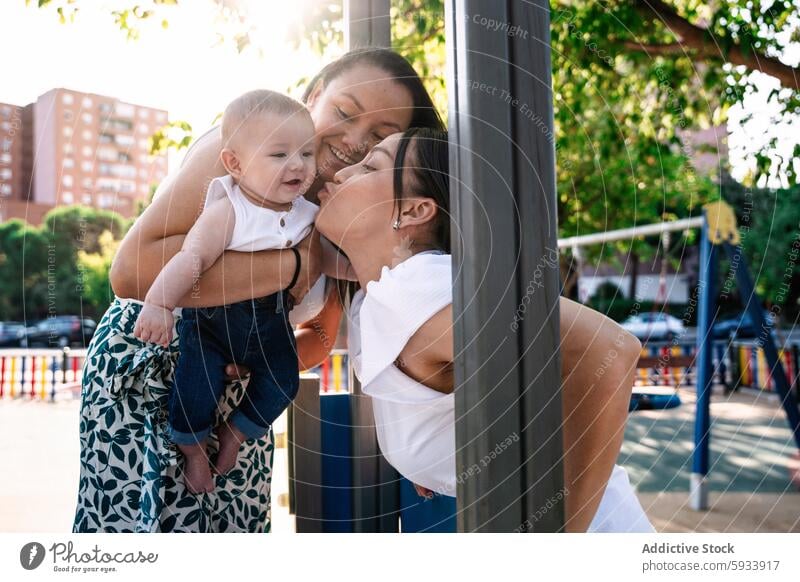 Mother and friend playfully kissing a baby at a sunny playground mother couple lesbian lgbt colombian joy outdoor sunlight happiness family bonding interaction