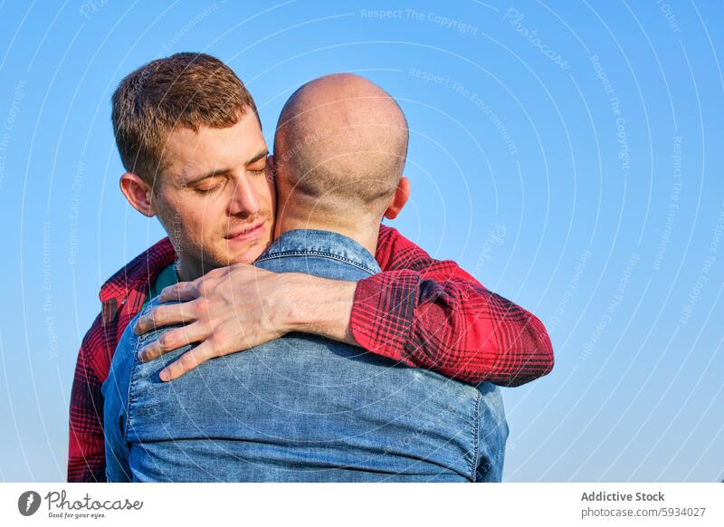 Emotional embrace between gay couple under clear sky emotional affection tender lgtbiq love relationship man hug comfort support denim plaid casual wear outdoor