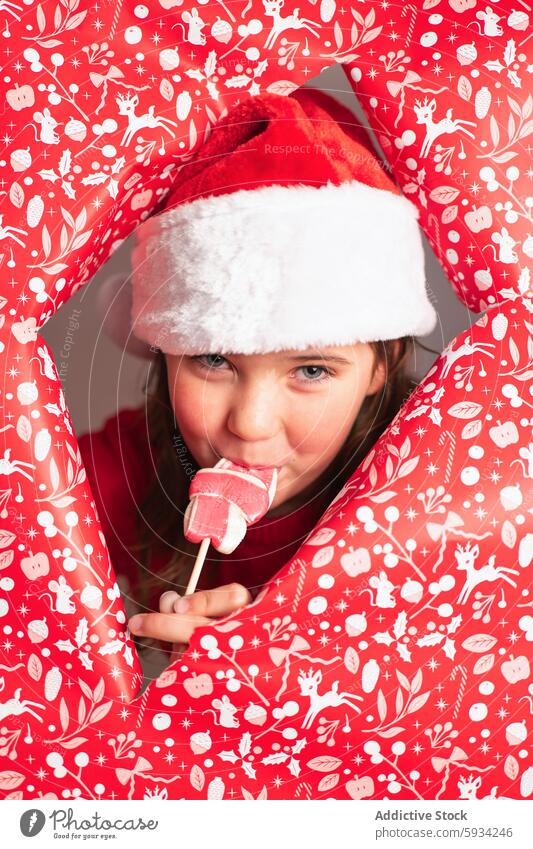 Young girl in Santa hat enjoying a lollipop in Christmas setting santa hat christmas fun playful red theme paper cutout hole savor vibrant festive holiday