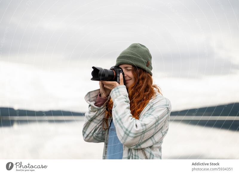 Woman taking photos at lake during camping trip woman camera photography landscape outdoor tranquil red hair beanie plaid jacket nature hobby scenery leisure