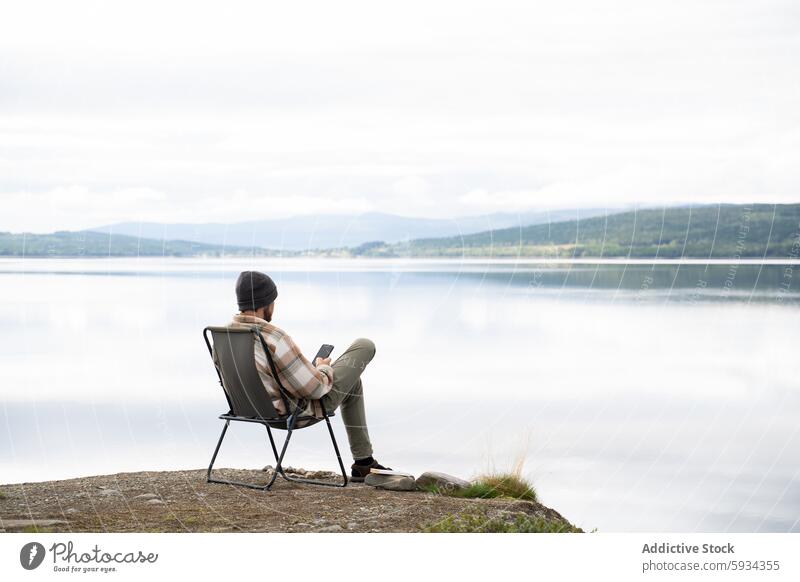 Man enjoying serene lakeside camping with a smartphone chair book man tranquil nature outdoor seated reading forest mountain mist solitude peaceful relaxation