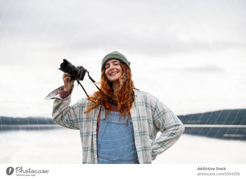 Smiling woman taking photos by a lake during camping camera photography outdoor nature cheerful smiling redhead beanie overalls relaxed hobby leisure joy