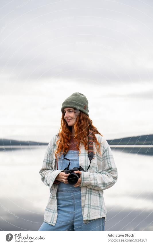 Young woman enjoying photography at a lakeside campsite camping nature outdoor casual redhead curly hair denim overalls beanie tranquil photographer female