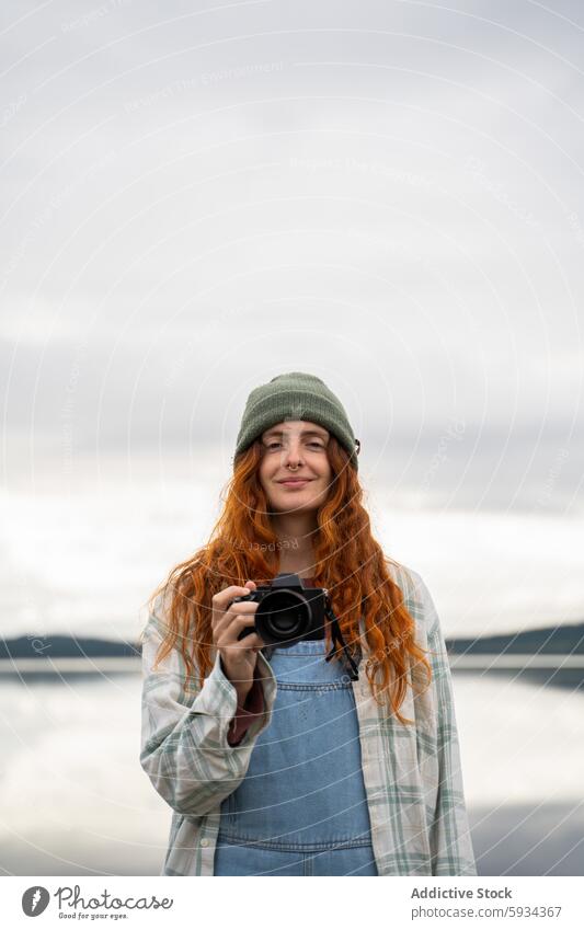 Red-haired woman with camera enjoying outdoor adventure red hair beanie denim overalls lake sky cloudy serene nature capture photography travel leisure hobby