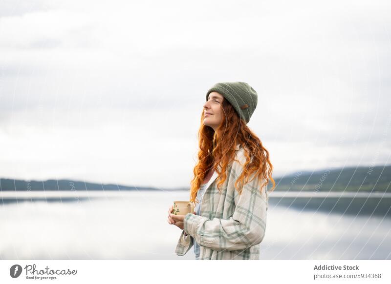 Serene young woman enjoying a lakeside camping morning outdoor relaxation serene beverage warm tranquil red-haired green beanie jacket plaid cozy nature