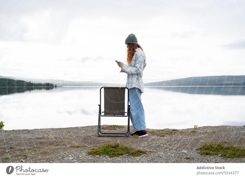 Young woman using smartphone by scenic lake while camping chair outdoor tranquil wilderness cloudy sky standing technology nature water morning leisure