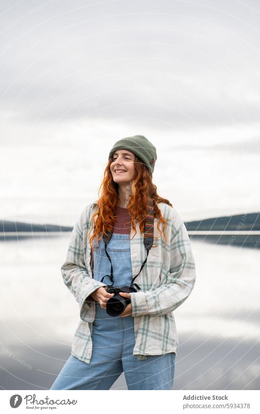 Red-haired woman enjoying nature while camping outdoor camera lake smile red hair cloudy casual attire joyful scenic lakeside adventure travel leisure hobby