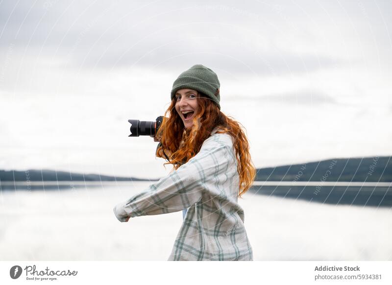 Happy woman taking photos by the lake during camping camera photography nature redhead cheerful outdoor happiness scenic view beanie plaid shirt green water