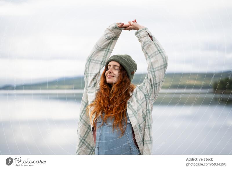 Woman enjoying a serene moment by the lake during camping woman morning nature outdoor stretch tranquility peaceful flannel shirt beanie denim overalls