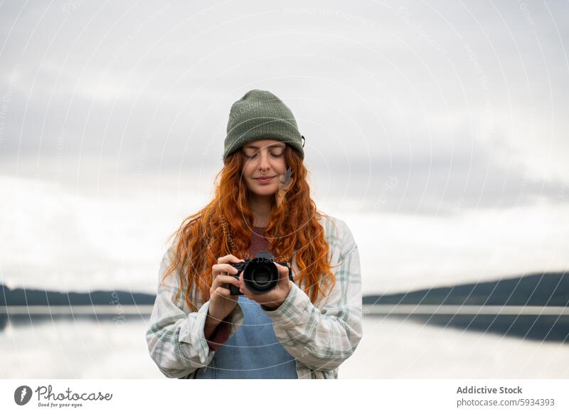 Young woman enjoys photography by lakeside camera beanie red hair camping serene nature peaceful outdoors plaid shirt solitude calm water landscape leisure