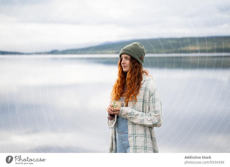 Serene woman enjoying coffee by the lakeside during camping redhead beanie serenity reflection morning outdoor nature peaceful plaid shirt denim landscape