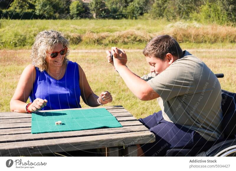 Boy in wheelchair playing dice game with woman outdoors boy cerebral palsy smile table field sky green blue interaction health disability care affection summer