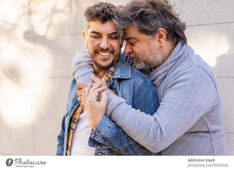 Gay couple sharing a tender embrace outdoors gay love affectionate joyful relationship happiness urban casual warm sweater denim jacket smiling beard grey hair