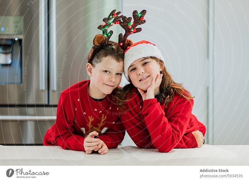 Twin siblings in festive outfits celebrating Christmas together twin boy girl christmas sweater reindeer antler santa hat smile joyful pose modern kitchen