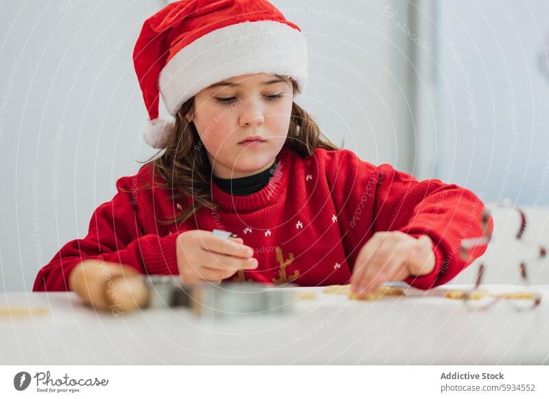 Young girl baking cookies in a festive Christmas outfit christmas holiday santa hat red sweater kitchen activity concentration cooking youth child seasonal