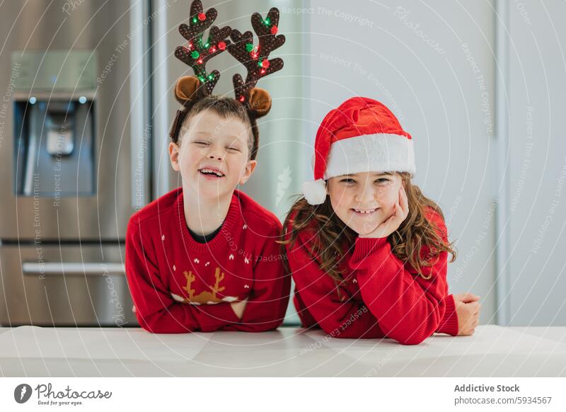 Twins dressed in Christmas outfits in a festive kitchen twin boy girl christmas laugh reindeer santa hat red sweater holiday decoration modern sibling joy