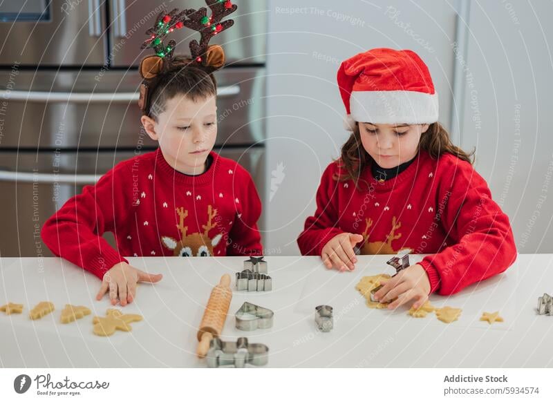 Twins baking holiday cookies in festive outfits twin child christmas sweater hat kitchen modern concentrate cheerful seasonal scene cutting decoration reindeer