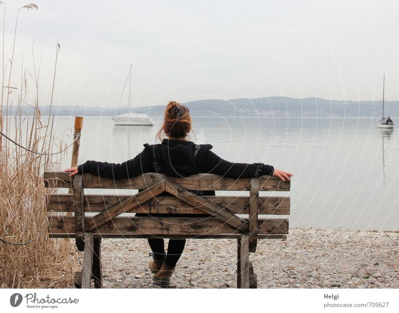 Rear view of a woman with pinned up brunette hair and dark jacket sitting on a wooden bench and looking at a lake in the fog Well-being Relaxation Calm