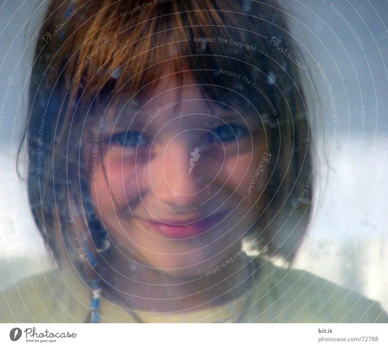 Cute, sweet, cute, smiling, little, dreamy, sweet, sweet girl with pony, looks cheekily, through a dirty pane of glass into the camera and observes, curious, his counterpart. The window is smeared with dirt and drops.