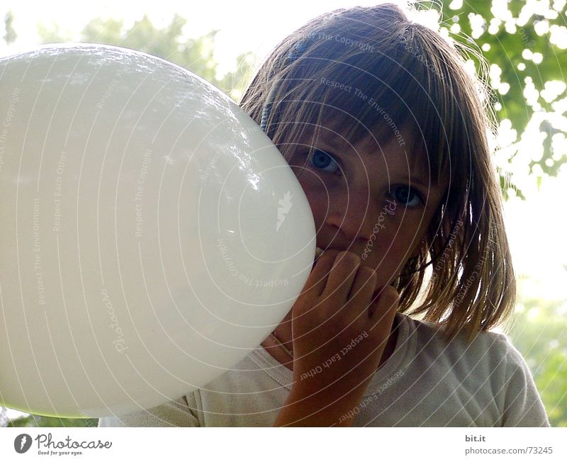 Thoughtful, dreamy, sweet girl with a white balloon in her hand in the garden, looks into the camera. Dear birthday girl outside in the nature is happy about a gift balloon for her party, holds it tight and puts it in her mouth.